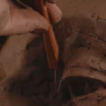 A hand sculpts clay using a wooden carving tool to shape a face.